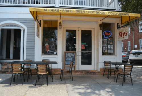 A restaurant exterior with outdoor seating, featuring a sign for sliders, fries, beer, hot dogs, and cold drinks.