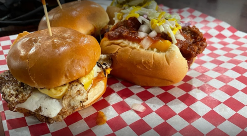 Two sandwiches on a red and white checkered paper: one with fried chicken and egg, the other with chili and toppings.
