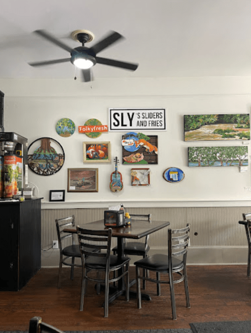 A cozy restaurant interior with a ceiling fan, tables, and colorful wall art, including a sign for "Sly's Sliders and Fries."