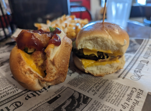A close-up of two sandwiches on newspaper: one with a hot dog topped with ketchup and pickles, the other a cheeseburger.
