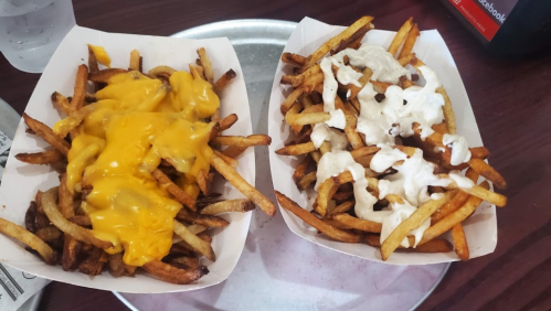 Two trays of French fries: one topped with melted cheese and the other with a creamy white sauce.