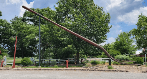 A large, artistic fork sculpture stands in a grassy area with trees and a blue sky in the background.