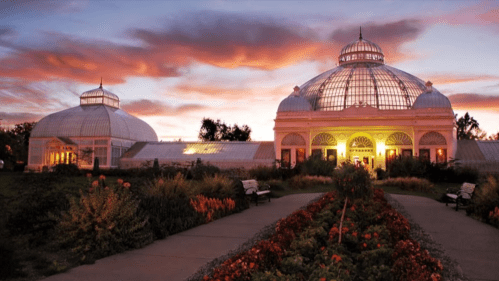 A beautiful sunset illuminates a botanical conservatory with glass domes, surrounded by colorful flowers and pathways.