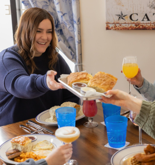 A woman smiles while serving biscuits at a table with drinks and plates of food, enjoying a meal with friends.