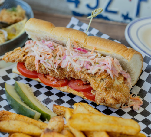 A crispy fried fish sandwich with coleslaw, tomatoes, pickles, and fries on the side, served on a checkered paper.