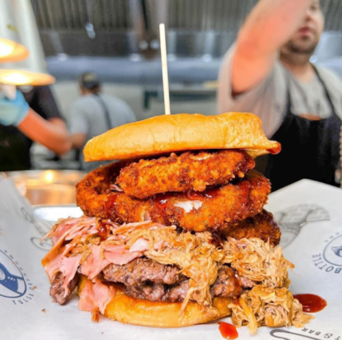 A towering burger with pulled pork, onion rings, and a beef patty, served on a bun with barbecue sauce.