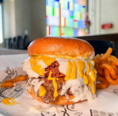 A juicy burger topped with melted cheese and creamy sauce, served with curly fries in a vibrant restaurant setting.