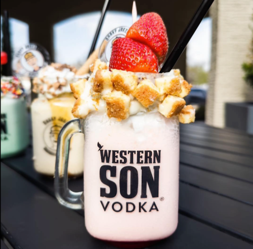 A creamy pink milkshake topped with whipped cream, strawberries, and cookie pieces in a glass with "WESTERN SON VODKA" logo.