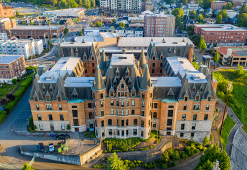Aerial view of a large, historic building with turrets, surrounded by modern buildings and greenery.