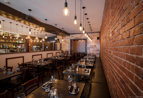 A cozy restaurant interior with wooden tables, chairs, and warm lighting against a brick wall.
