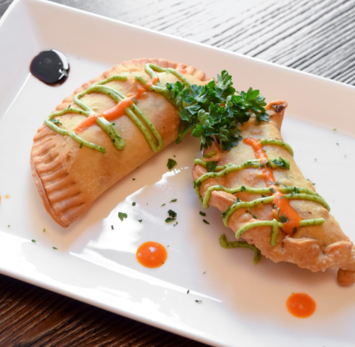 Two golden-brown empanadas drizzled with green and red sauces, garnished with parsley on a white plate.