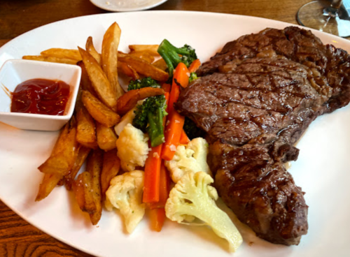 A plate featuring a grilled steak, crispy fries, and a mix of steamed vegetables with a small bowl of ketchup.