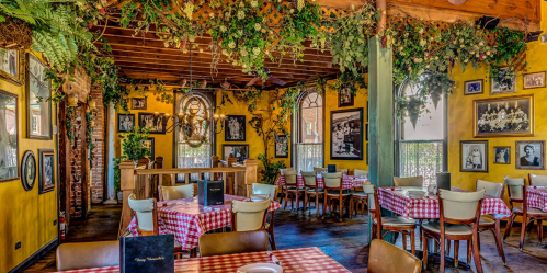 Cozy restaurant interior with checkered tablecloths, greenery, and framed photos on yellow walls. Warm, inviting atmosphere.