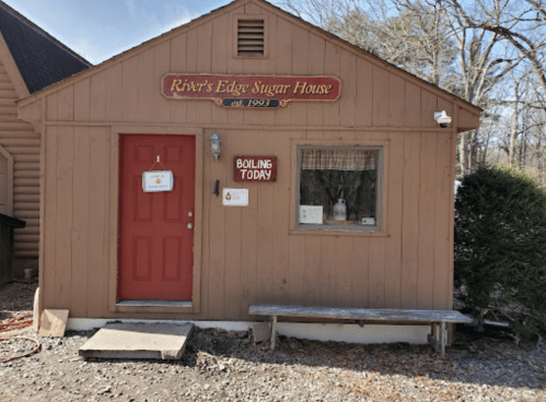 A small, rustic sugar house with a red door and sign reading "River's Edge Sugar House" and "Boiling Today."