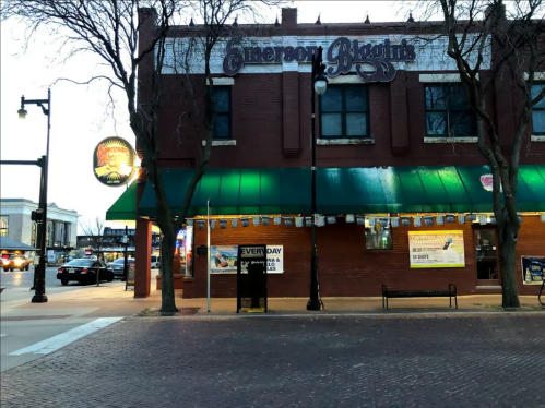 A brick building with a green awning, featuring a sign for Emerson Biggins and outdoor seating, in an urban setting.