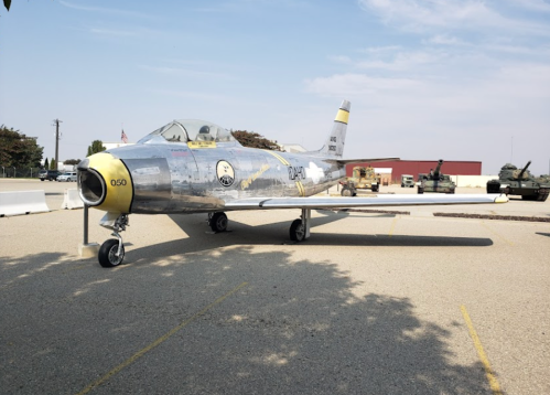 A vintage silver jet aircraft displayed outdoors, with military vehicles in the background and a clear sky above.