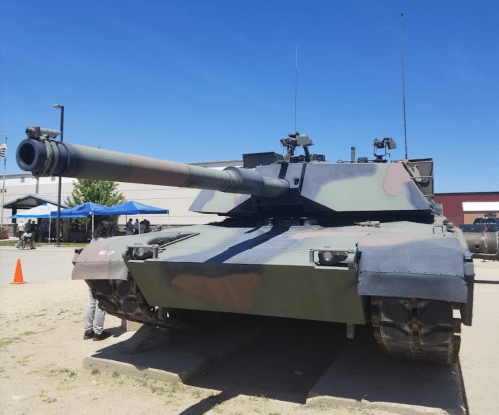 A military tank displayed outdoors, featuring a camouflage pattern and a long barrel, with people in the background.