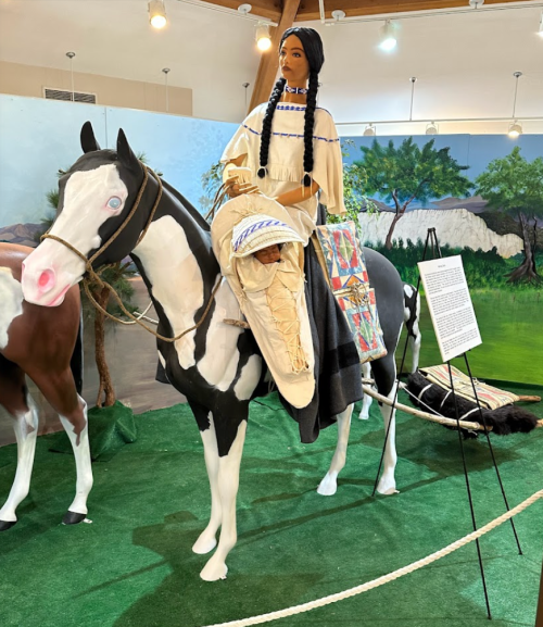 A life-sized mannequin of a woman in traditional attire riding a painted horse, displayed in an exhibit.