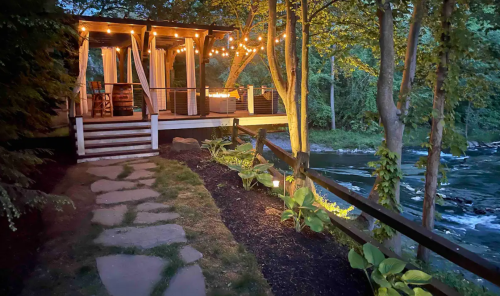 Cozy riverside cabin with string lights, surrounded by trees and a stone pathway leading to the water.