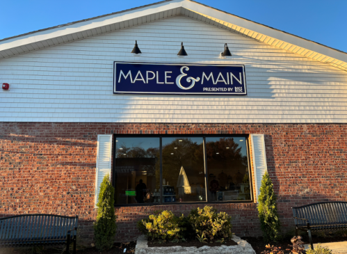 Exterior of a building with a sign that reads "Maple & Main" and large windows, surrounded by greenery and benches.