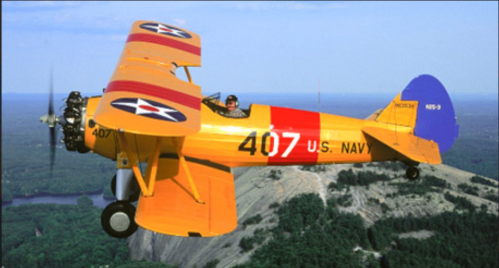 A bright yellow biplane with military markings flying over a mountainous landscape.