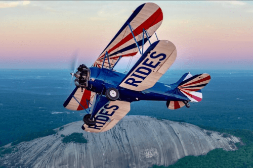 A blue biplane with "RIDES" on its wings flying over a large rock formation at sunset.