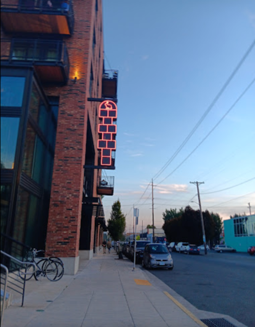 A brick building with a neon sign, street view showing parked cars and bicycles along the sidewalk under a blue sky.