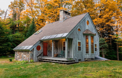 A cozy wooden cabin surrounded by vibrant autumn foliage, featuring large windows and a stone chimney.
