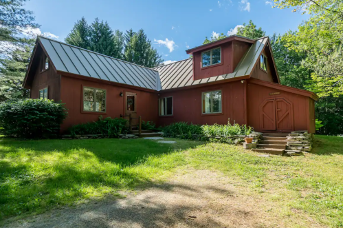 A red house with a metal roof surrounded by greenery and trees on a sunny day.