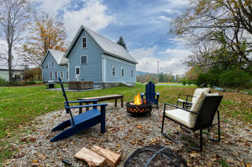 A cozy outdoor fire pit area with blue chairs, a fire burning, and a light blue house in the background surrounded by autumn leaves.