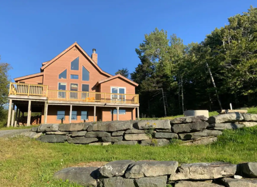 A large wooden house with a deck, surrounded by trees and a stone wall, under a clear blue sky.