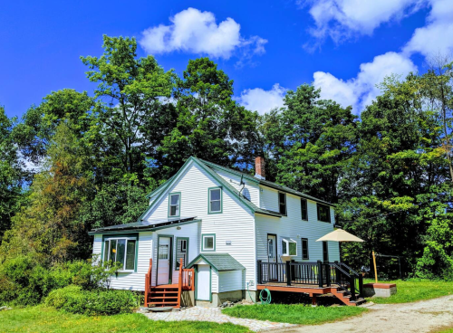 A white two-story house surrounded by trees, featuring a deck and a small outdoor area under a blue sky.
