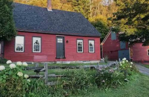 A red farmhouse with a black door, surrounded by greenery and flowers, set against a backdrop of trees.