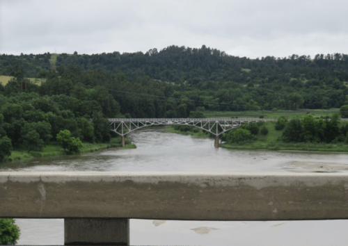 A metal bridge spans a river, surrounded by lush greenery and hills under a cloudy sky.