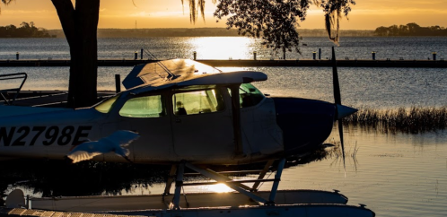 A seaplane docked by a calm lake at sunset, with reflections on the water and trees in the background.