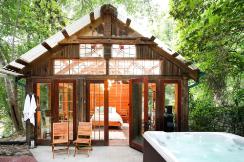 A rustic cabin with large glass doors, surrounded by trees, featuring a hot tub and wooden chairs on a patio.