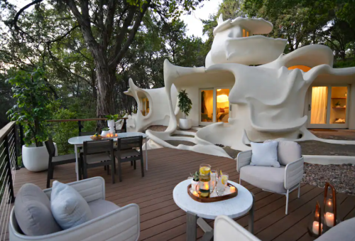 A unique, sculptural white house surrounded by trees, featuring a wooden deck with outdoor seating and decor.