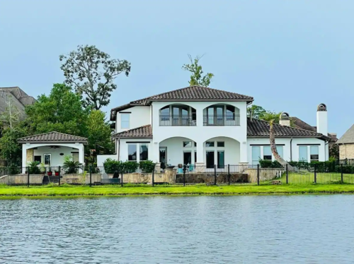 A modern two-story house with a tiled roof, surrounded by greenery, located by a calm lake.