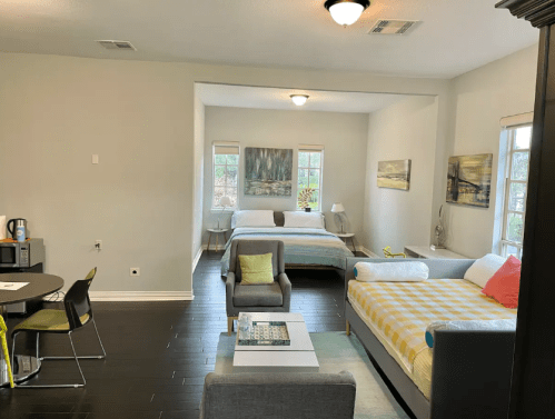 A cozy room with two beds, a small dining table, and artwork on the walls, featuring natural light from windows.