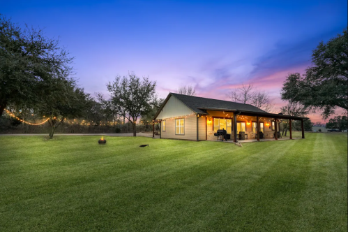 A cozy house with warm lights, surrounded by green grass and trees, under a colorful sunset sky.