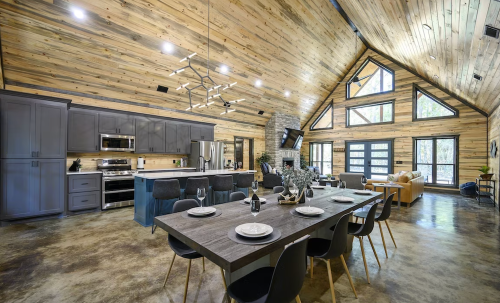 Modern kitchen and dining area with wooden walls, high ceilings, and large windows, featuring a stylish table and seating.