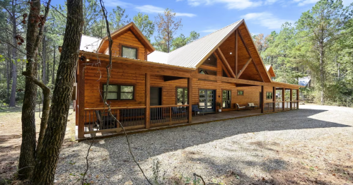 A spacious wooden cabin surrounded by trees, featuring a large porch and a sloped roof under a clear blue sky.