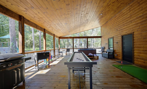 A spacious wooden patio with a shuffleboard table, grill, and views of trees through large windows.
