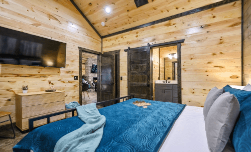 Cozy bedroom with wooden walls, a blue quilted bedspread, TV, and a chess set on the bed. Modern rustic decor.