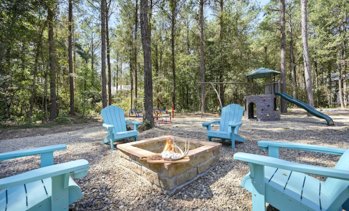 A cozy outdoor space with blue chairs around a fire pit, surrounded by trees and a playground in the background.