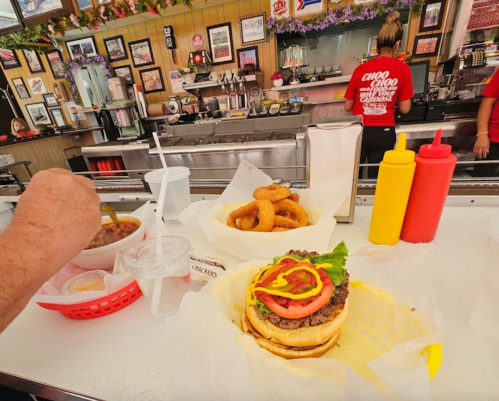 A diner table with a burger topped with lettuce and tomato, onion rings, chili, and condiments in a retro-style restaurant.