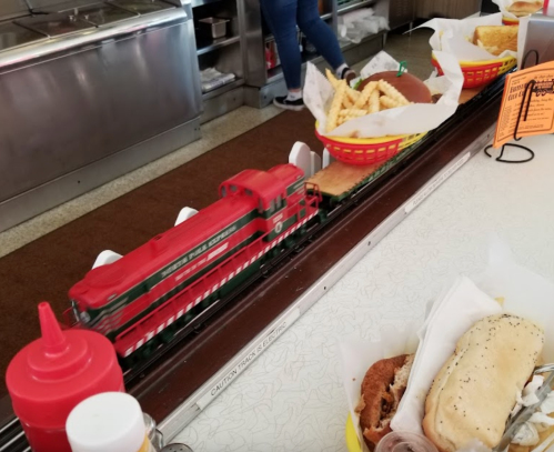 A toy train delivers baskets of fries and sandwiches along a diner counter, with condiments in the foreground.