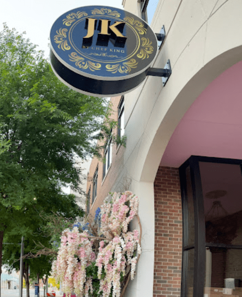 Sign for "JK by Chef King" with floral decorations outside a building, surrounded by greenery.