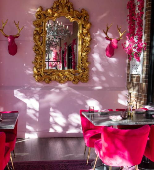 A stylish restaurant interior with pink walls, ornate mirror, and decorative antlers, featuring pink chairs and tables.