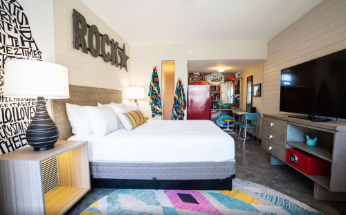 A modern hotel room featuring a bed, colorful decor, a TV, and a kitchenette area with retro-style furniture.